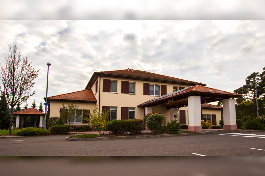 Two-story cream-colored building with red shutters and terracotta roof, cloudy sky, parking area in front.
