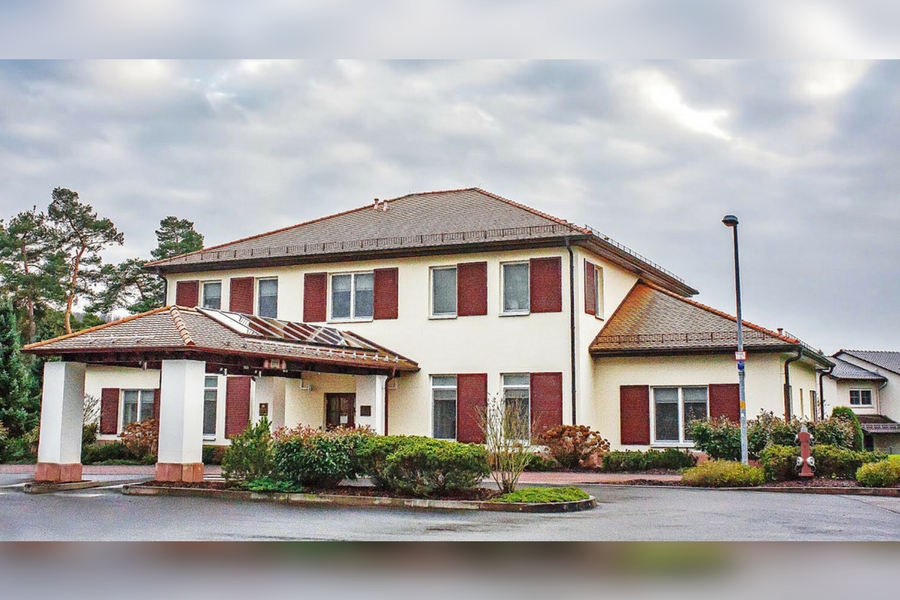 Large cream building with dark red shutters and tiled roof, landscaped bushes, cloudy sky, parking lot in front.